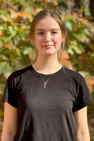 This is a portrait of a young woman with short hair wearing a black t-shirt standing outdoors in front of autumn foliage.