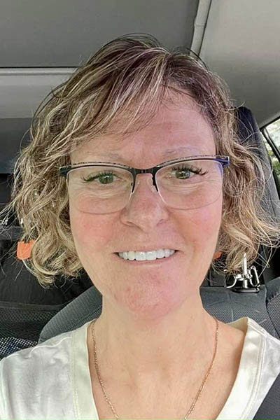 A smiling woman with short hair wearing glasses and a blue shirt, taken from inside a vehicle.
