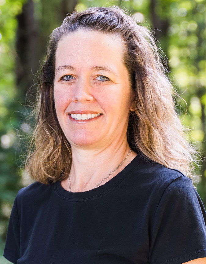 The image shows a smiling woman with short hair wearing a black shirt and standing outdoors against a natural background.