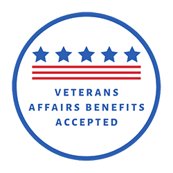 The image features a circular logo with a blue background containing a white border. At the center, there is an emblem consisting of five stars arranged in a horizontal line above a red stripe. Below the stripe, there is text that reads  VETERANS AFFAIRS BENEFITS APPROVED  in capital letters. The logo has a badge-like design and appears to be associated with a specific organization or certification related to veterans  affairs benefits.
