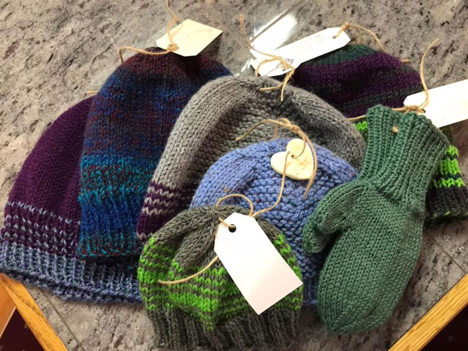 The image displays a collection of handcrafted knit hats with various patterns and colors, presented on a table with price tags attached to them.