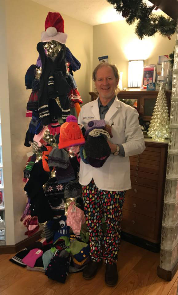 Man standing beside Christmas tree adorned with various hats and holiday decorations.