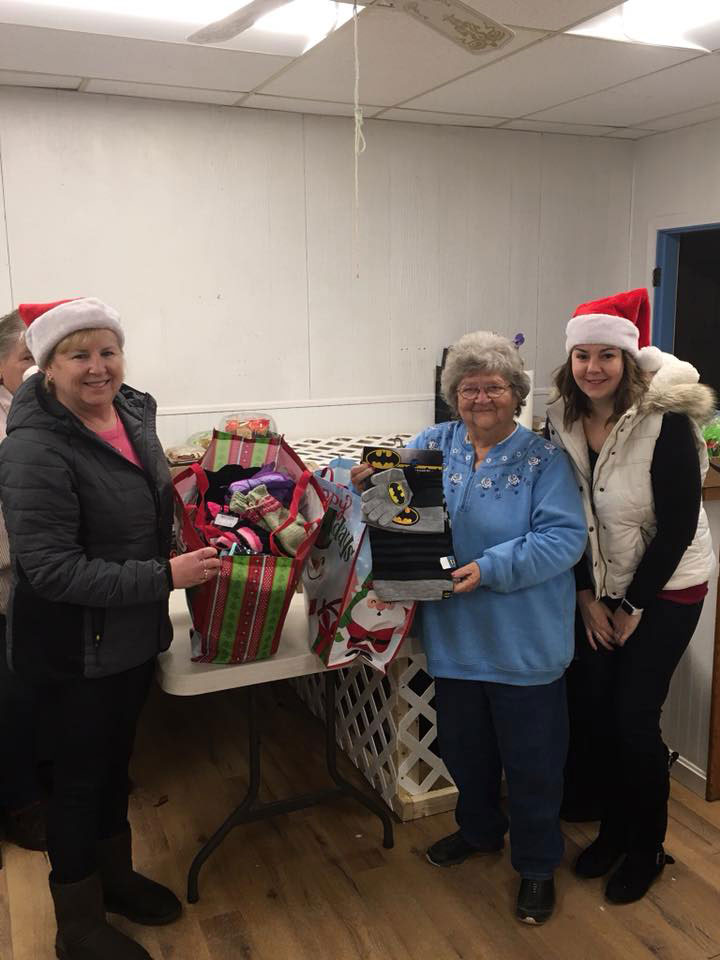 The image shows three individuals standing behind a table with gifts on it, holding a trophy with a plaque, posing for a photo with smiles, wearing festive holiday-themed attire, and they are indoors in a room with a Christmas decoration hanging from the ceiling.