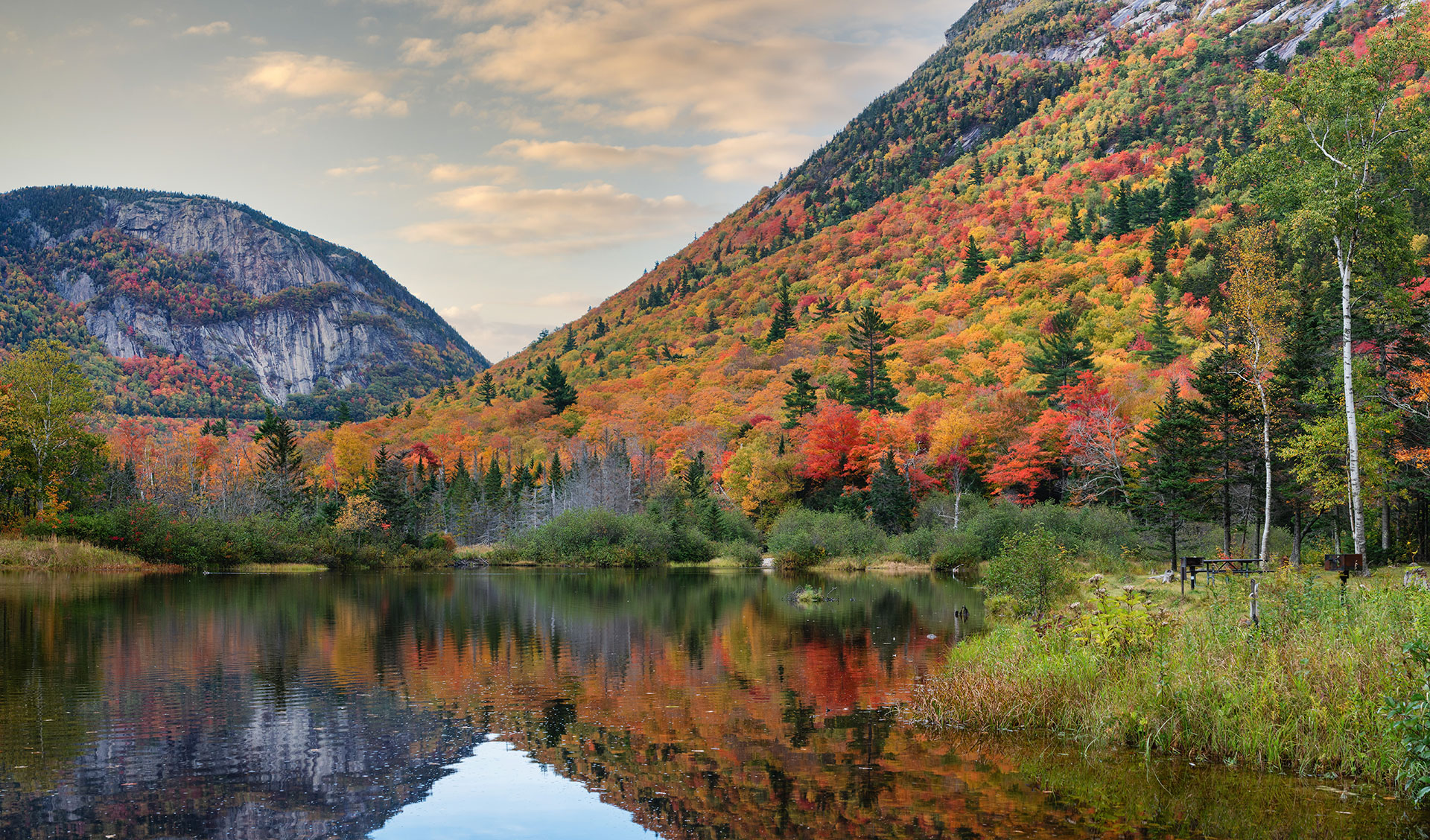 The image shows a serene autumn landscape with a vibrant red foliage-covered hillside, a calm lake reflecting the surrounding colors, and a clear blue sky above.