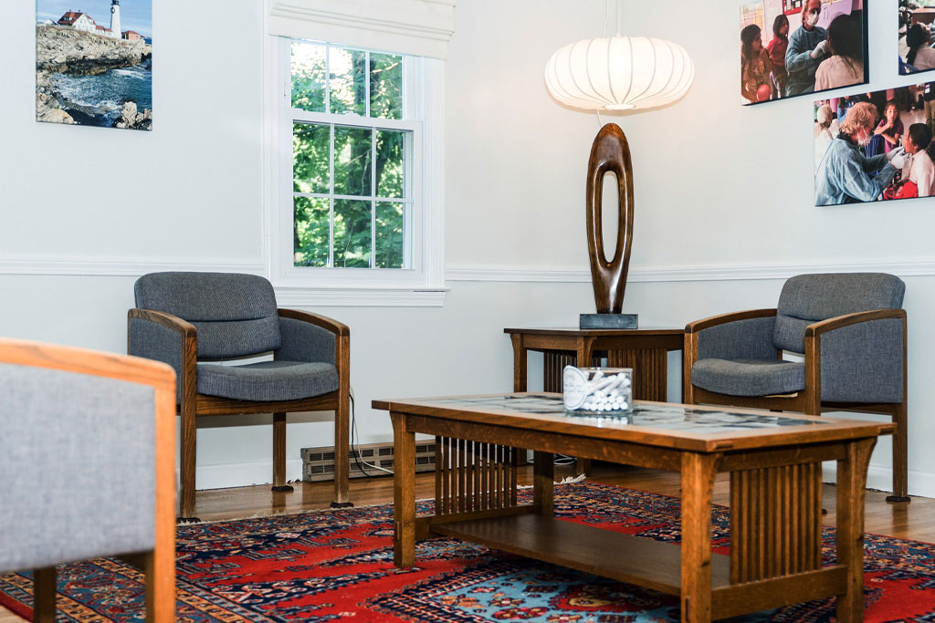 The image shows a well-lit room with two chairs facing each other around a small wooden table, a large abstract wall art, a modern pendant light fixture hanging from the ceiling, and a framed photograph on the wall.