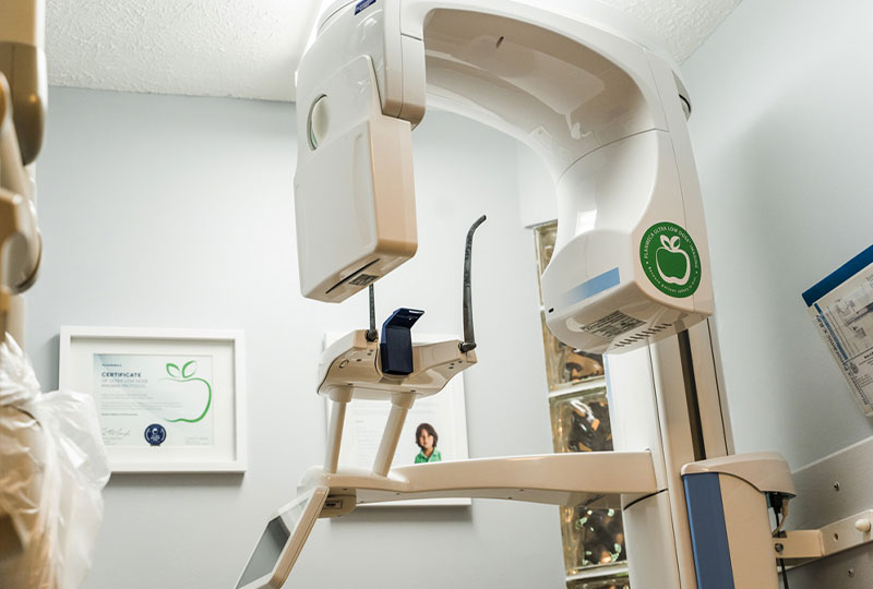 The image shows an interior space with medical equipment, including a large dental chair, a panoramic X-ray machine, and a wall-mounted monitor displaying an X-ray image. There are posters and a framed certificate on the wall behind the equipment. A person is partially visible at the bottom of the frame, suggesting the presence of a medical professional or patient in the room.
