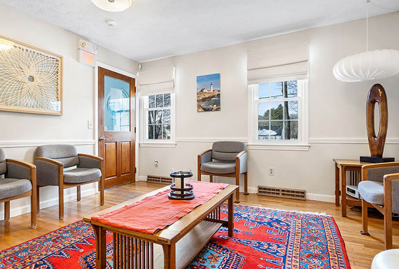 The image shows a well-lit living room with modern furnishings, including a sofa, chairs, a coffee table, and decorative elements such as artwork on the walls and a vase on the table. The space has a cozy atmosphere with a view of a window revealing an outdoor setting.