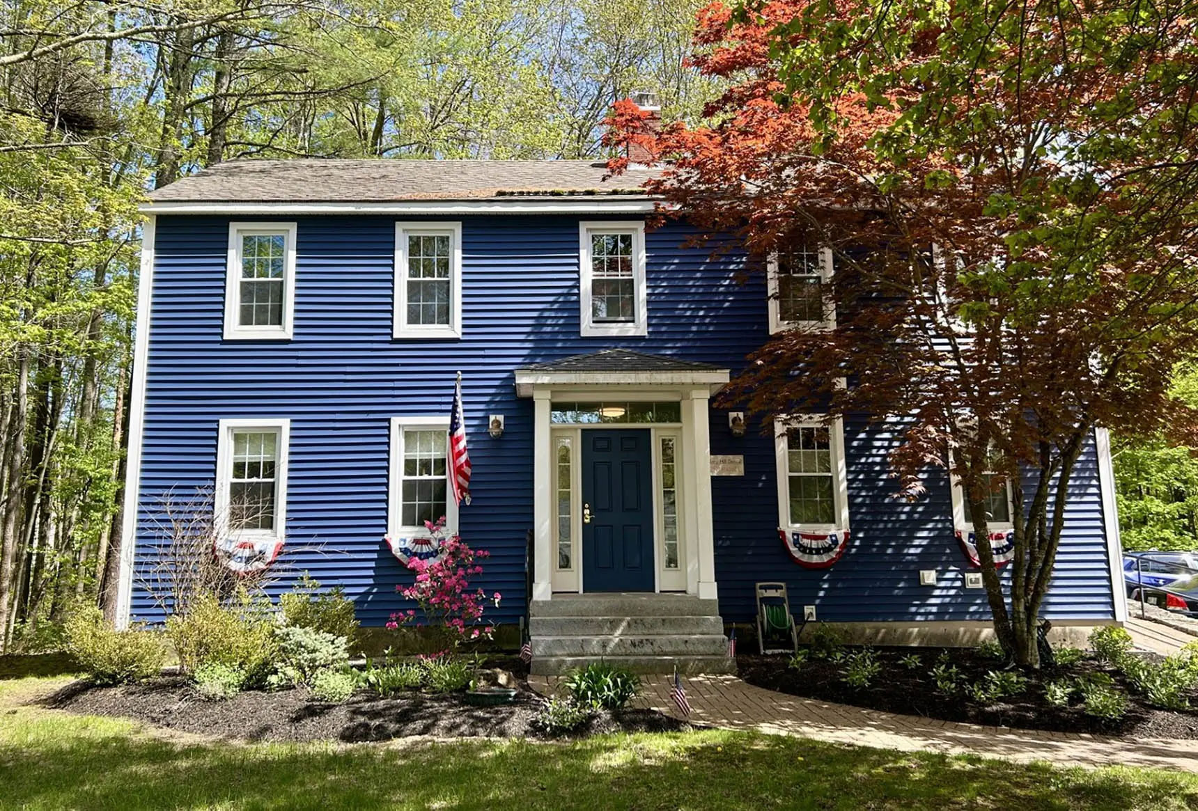 The image depicts a two-story blue house with white trim, a front porch, and American flags hanging on either side of the door. The house is surrounded by a well-manicured lawn with a few trees in the background, and there are red autumn leaves visible on some branches.