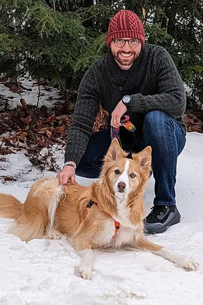 A man kneeling on snow next to a brown dog with an orange harness.