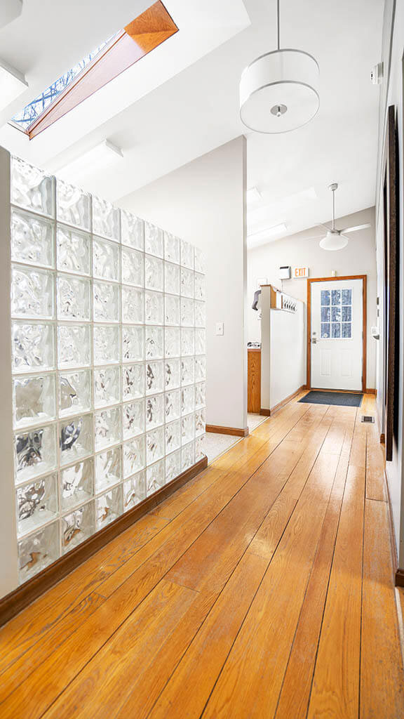 The image shows an interior hallway with hardwood flooring, white wall tiles, and shelves displaying various items on both sides.