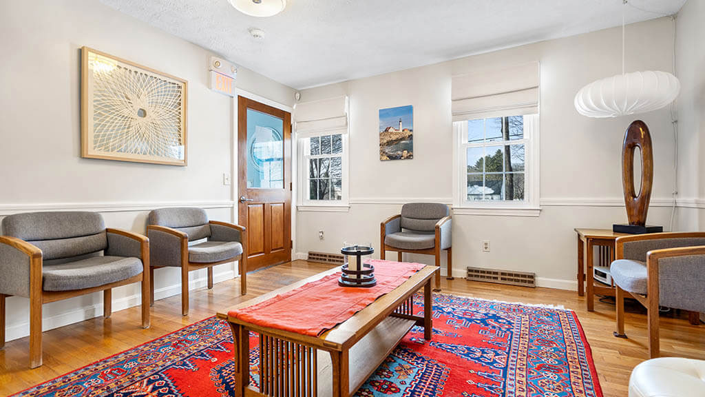 The image shows an interior space with a living room area featuring a red rug, a wooden coffee table, and chairs, alongside a hallway leading to another room with a glimpse of a couch and artwork on the wall.
