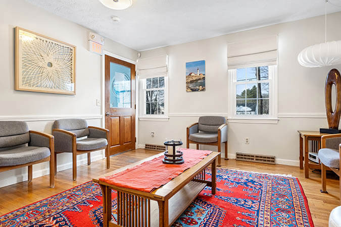 The image depicts a well-lit living room with modern furniture, including a sofa and chairs, a red area rug, and artwork on the walls.