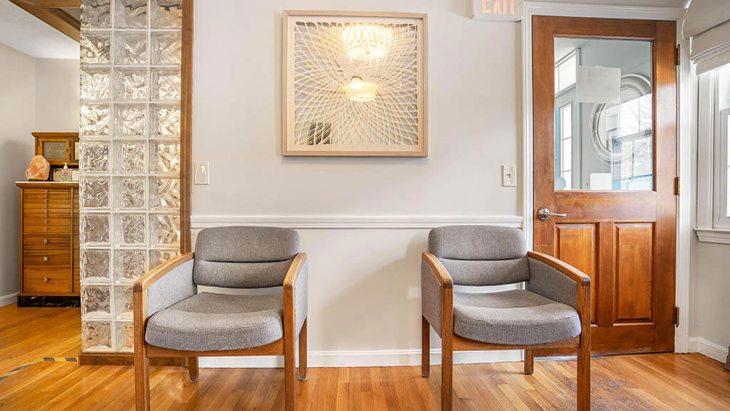 The image shows a well-lit waiting room with modern furniture, including two chairs and a bench, positioned against a wall with a decorative panel and framed artwork above it.