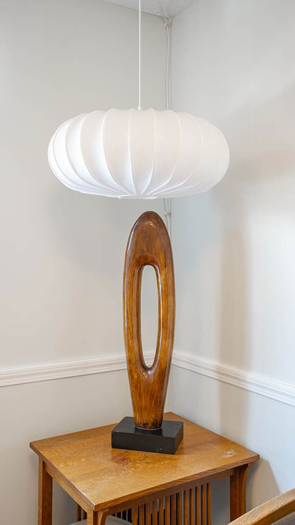 The image shows a modern interior with a large, abstract sculpture on a pedestal next to a wall-mounted light fixture shaped like a flower or sunburst design, set against a backdrop of a simple white wall and a piece of furniture that appears to be a table or sideboard.