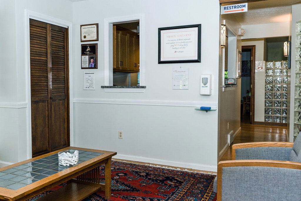 The image shows two photos of an interior space, likely an office or waiting area, with a wall-mounted sign, a reception desk, a carpeted floor, and various pieces of furniture including chairs, tables, and cabinets.
