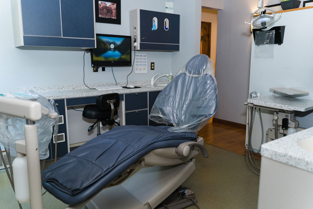 The image shows a dental office interior with a chair covered in plastic, a computer monitor displaying an image, a sink, and various dental equipment.