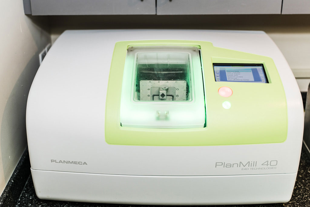 The image shows a modern laboratory device with a digital interface, labeled 'PlanMill 3D,' which appears to be a piece of equipment for 3D printing or scanning. It's placed on a table in an indoor setting, possibly a lab or office environment.