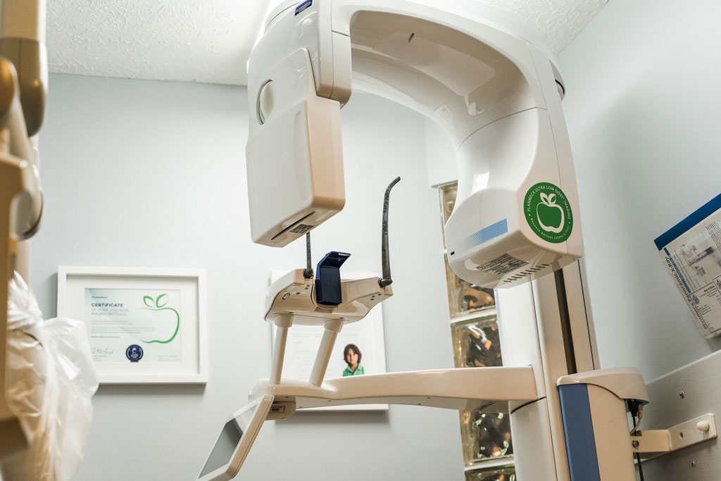 The image shows a modern dental practice with a large, advanced dental chair equipped with a digital imaging system and a ceiling-mounted CAT scan machine, set against a white wall with a framed picture hanging above the chair.