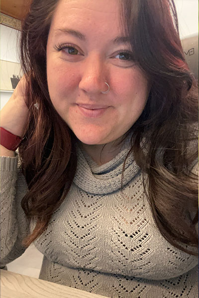 A woman with long hair smiling at the camera, wearing a grey sweater and sitting in an office setting.